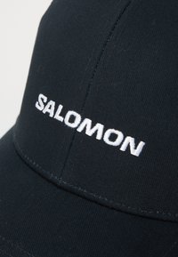 Melna cepure, kas izgatavota no izturīga auduma, ar balto izšuvumu "SALOMON" logotipu priekšpusē. Gluda tekstūra ar strukturētu dizainu.