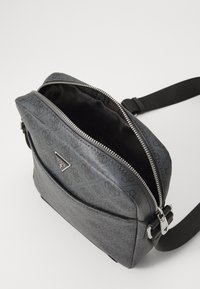 Bolso de mano gris hecho de cuero sintético texturizado, con cierre de cremallera, correa ajustable y un destacado logo en la parte delantera.