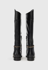 Juodos odinės kelio ilgio botas su elegantišku dizainu, smailiais viršūnėmis, užtrauktuko detale šone ir auksine grandine aplink kulkšnį.
