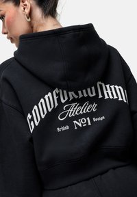 Felpa nera con una grande scritta grafica bianca sul retro, che recita "Goodforanthing Atelier No.1 British Design." Materiale in cotone, texture morbida.