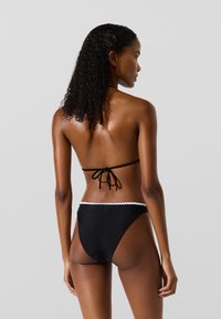 Čierne bikini s viazaným halter topom a vysokými nohavičkami s bielym vlnkovaným lemovaním. Hladká textúra s minimálnym zakrytím.