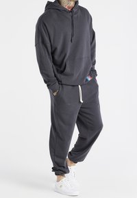 Felpa con cappuccio grigio scuro e pantaloni della tuta abbinati in tessuto morbido. Presenta una tasca frontale e un cappuccio con coulisse regolabile. Indossati con sneakers bianche.