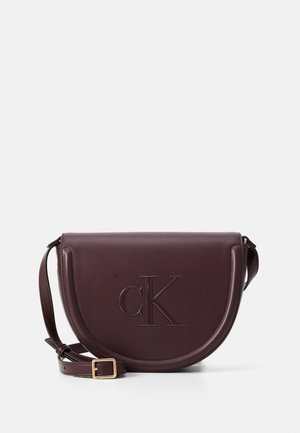 Bolso bandolera de cuero color púrpura oscuro en forma de media luna, con correa ajustable y logo “cK” en relieve en la solapa delantera.