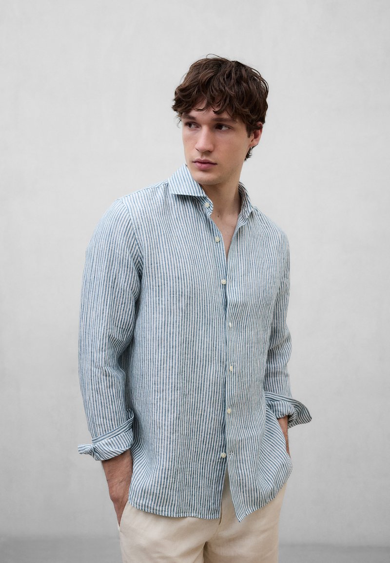 Joven con cabello castaño ondulado, vestido con una camisa de rayas azules y blancas abotonada y pantalones beige, mirando hacia un lado con las manos en los bolsillos.