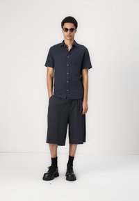 Chemise à manches courtes bleu marine à boutons avec un tissu texturé et une poche, associée à un short noir. Le mannequin porte des lunettes de soleil noires et des chaussures noires imposantes.