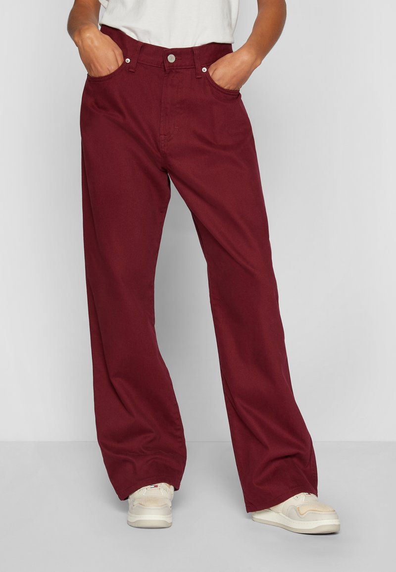 Pantalones de pierna ancha burdeos hechos de algodón, con una cintura alta, dos bolsillos frontales y un cierre de botón. Combinados con zapatillas de color claro.