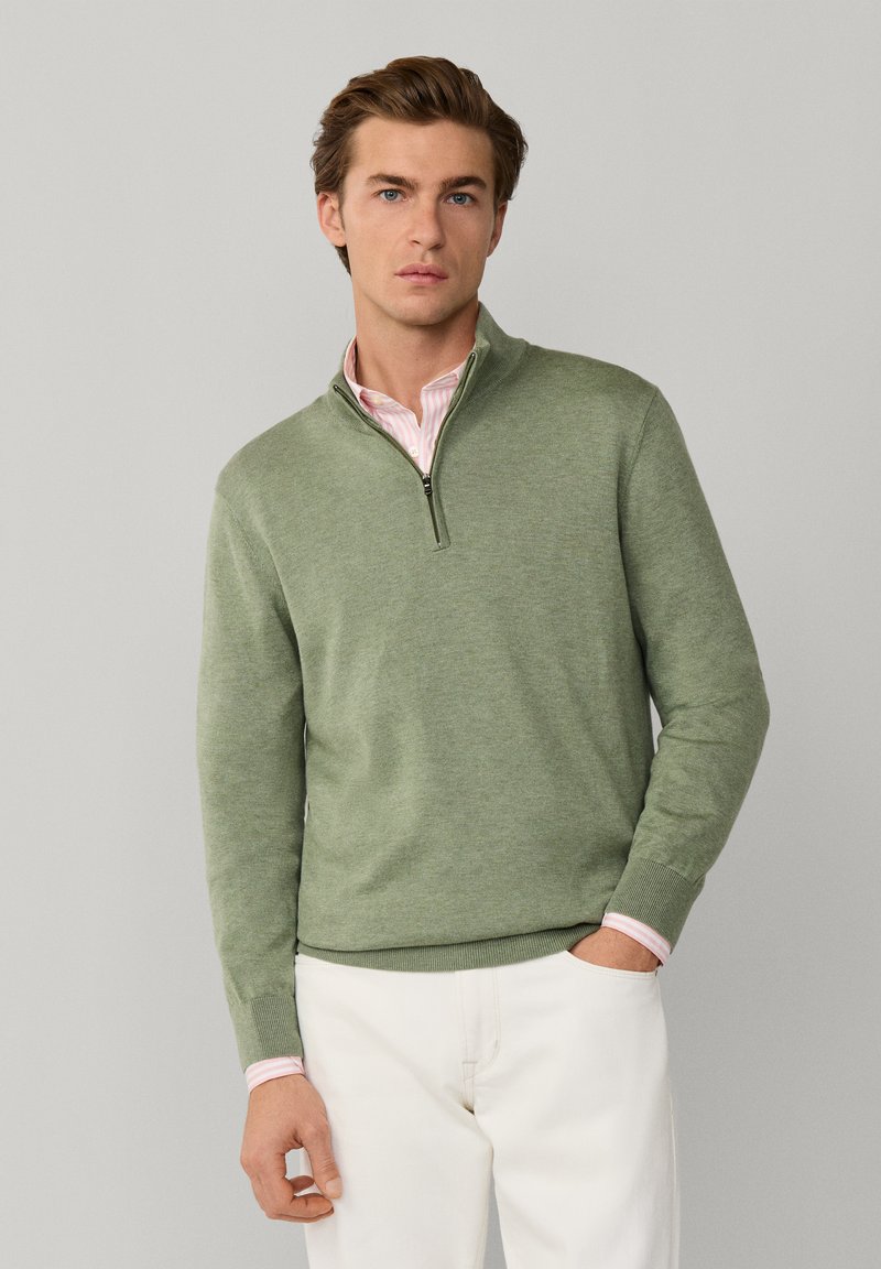 Młody mężczyzna o jasnobrązowych włosach, ubrany w zielony sweter z zamkiem do połowy, nałożony na różową koszulę i białe spodnie, stoi na jednolitym tle.