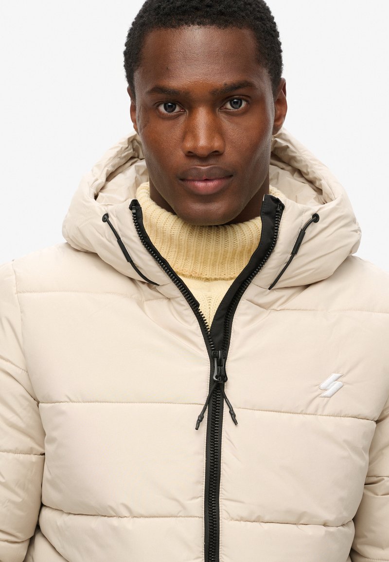 Beige Daunenjacke mit Kapuze, ausgestattet mit einem Reißverschluss und schwarzen Akzenten; getragen über einem cremefarbenen gestrickten Pullover mit hohem Kragen.