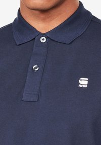 Homme portant un polo bleu marine avec deux boutons et un logo blanc G-Star Raw sur la poitrine gauche.