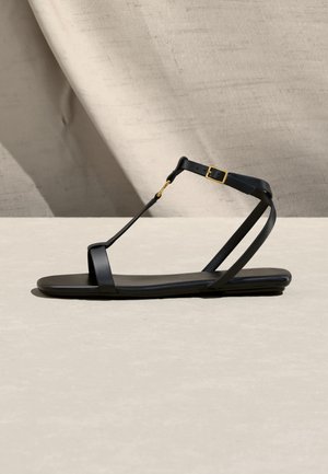 Sandale plate noire minimaliste avec fines lanières, boucle à la cheville et ferrures dorées, présentée sur un fond et une surface neutres.