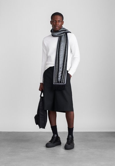 Fekete és szürke csíkos sál fröccsöntéssel, fehér pulóver fölé viselve. A modellen fekete nadrág és cipő is van, és egy sötét táskát tart.