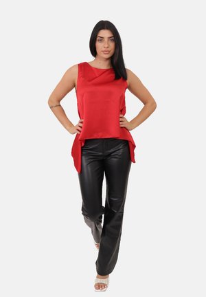 Kobieta z długimi ciemnymi włosami, ubrana w bezrękawnik w kolorze czerwonym, czarne skórzane spodnie, białe sandały na obcasie, stoi z rękami na biodrach na tle białym.