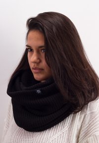 Black knitted infinity scarf with a textured pattern, featuring a small blue logo tag. Worn draped loosely around the neck.