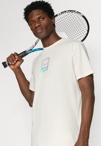 Κρέμα ριγέ μπλούζα φτιαγμένη από απαλό υλικό, με πολύχρωμο σχέδιο λογότυπου στο στήθος. Ο φορέας κρατάει μία ρακέτα με το ένα χέρι.