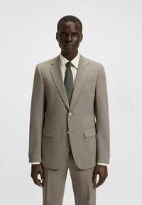 Lichtbruine, gestructureerde suit met een figuurlijke pasvorm, met twee voorzakken en een enkele knoop. Gecombineerd met een crèmekleurig shirt en een donkergroene stippenstrik.