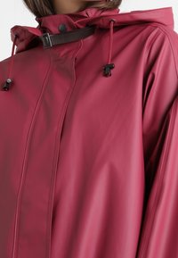Czerwona wodoodporna kurtka z kapturem, z zamkiem błyskawicznym z przodu, czarnymi akcentami i regulowanymi sznurkami. Gładka, błyszcząca faktura.