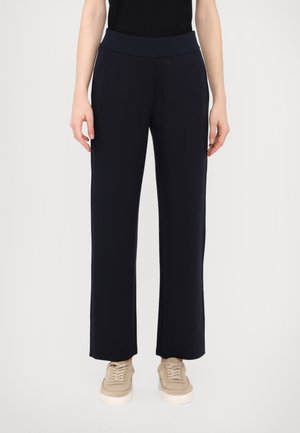 Person trägt schwarze, weit geschnittene Hosen und beige Sneakers, steht vor hellem, einfarbigem Hintergrund.