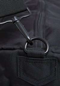 Black duffel bag detail featuring a metal clip and ring, with textured fabric and reinforced stitching on the handle attachment.