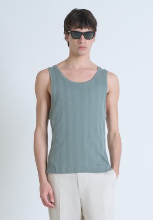Hellgrünes geripptes Tanktop mit rundem Halsausschnitt und breiten Trägern; getragen mit beigen, lockeren Hosen. Schwarze Sonnenbrillen ergänzen das Outfit.