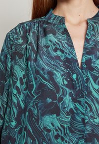 Chemise avec un décolleté en V profond, présentant un motif marbré dans des tons de vert et de noir. Le tissu a une texture lisse avec des accents froncés aux épaules.