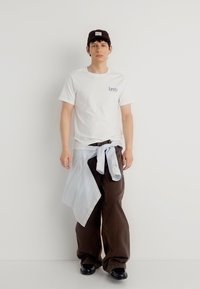 Νεαρός άνδρας που φοράει λευκό μπλουζάκι Levi's, καφέ φαρδιά παντελόνια, μαύρα παπούτσια, μαύρο καπέλο και ριγέ πουκάμισο δεμένο γύρω από τη μέση του.