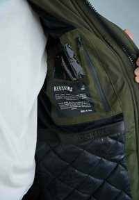 Veste en softshell olive foncé avec un intérieur matelassé noir. Présente une poche transparente et une étiquette de marque REDSKINS sur la doublure intérieure.