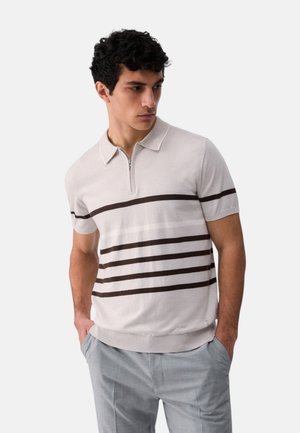 Giovane uomo che indossa una polo beige a maniche corte con righe orizzontali marrone scuro e pantaloni grigio chiaro, con le mani nelle tasche, guarda in basso a sinistra.