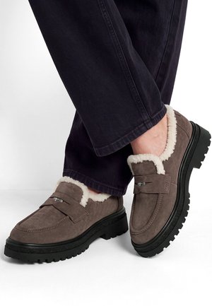Scarpe scamosciate marroni slip-on con fodera in pile bianco indossate con pantaloni neri, mostrate con gambe incrociate su sfondo bianco.