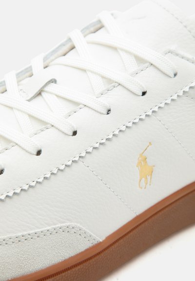 Λευκό δερμάτινο αθλητικό παπούτσι με λευκά κορδόνια, μπεζ σουέντ προστασία στα δάχτυλα, καφέ σόλα και χρυσό λογότυπο Polo στο πλάι.