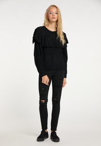 Fekete fodros pulóver texturált anyagból, fekete szakadt skinny farmerral és fekete sportcipővel párosítva. Egyszerű, monokróm öltözet.