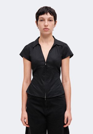 Jeune personne aux cheveux courts et foncés, portant un t-shirt noir ajusté à manches courtes avec fermeture éclair et un pantalon noir sur un fond uni.