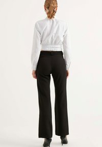 Vit kortärmad blus med rynkad rygg och långa ärmar, i kombination med tajta svarta byxor och svarta högklackade skor, sedd från baksidan.