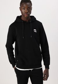 Zwarte hoodie van zacht materiaal, met een voorzak, een capuchon met trekkoord en een klein wit logo op de borst met tekst op de mouw.