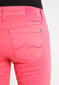 Różowe jeansy z denimu z tylną kieszenią ozdobioną przeszyciem w kształcie fali oraz małym logo marki na pasku od spodni.