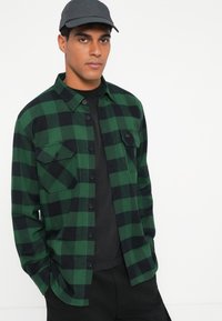Camicia di flanella a quadretti verde e nera con due tasche sul petto, bottoni neri e colletto, indossata sopra una maglietta nera.