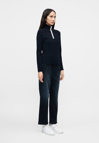 Γυναίκα όρθια, με μαύρο φούτερ με φερμουάρ, σκουρόχρωμο τζιν με φαρδιά μπατζάκια, λευκές κάλτσες και λευκά αθλητικά παπούτσια, μπροστά σε ένα απλό φωτεινό φόντο.