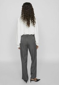 Blusa blanca de manga larga con puños fruncidos, combinada con pantalones grises de talle alto, de corte recto y textura sutil. Zapatos de tacón negros.