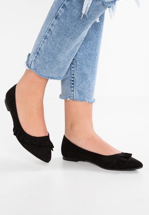 Voeten met zwarte platte schoenen met puntige neus en ruches, gecombineerd met lichtblauwe cropped jeans met rafelige zoom, op een witte achtergrond.