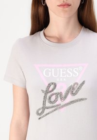 Svetlo šedé tričko s ružovým trojuholníkovým logom a textom "GUESS". Kryštálové akcenty tvoria slovo "Láska" v kurzíve naprieč prednou časťou.