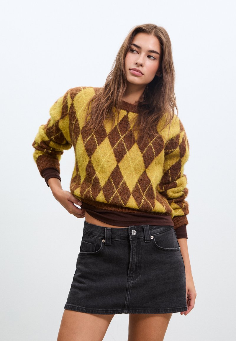 Femme portant un pull à losanges jaune et marron et une mini-jupe en jean noire, debout avec une main soulevant l'ourlet du pull.