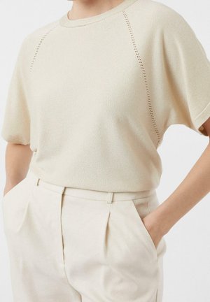 Person trägt ein hellbeiges kurzärmeliges Strickoberteil mit perforierten Details und passende hoch taillierte plissierte Hose mit den Händen in den Taschen.