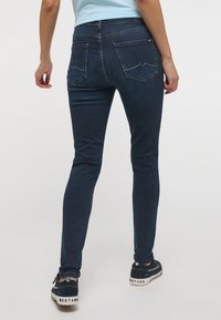 Jeans skinny azul oscuro con una textura suave, desvanecimiento sutil, dos bolsillos traseros con un diseño curvado, combinados con zapatillas oscuras.