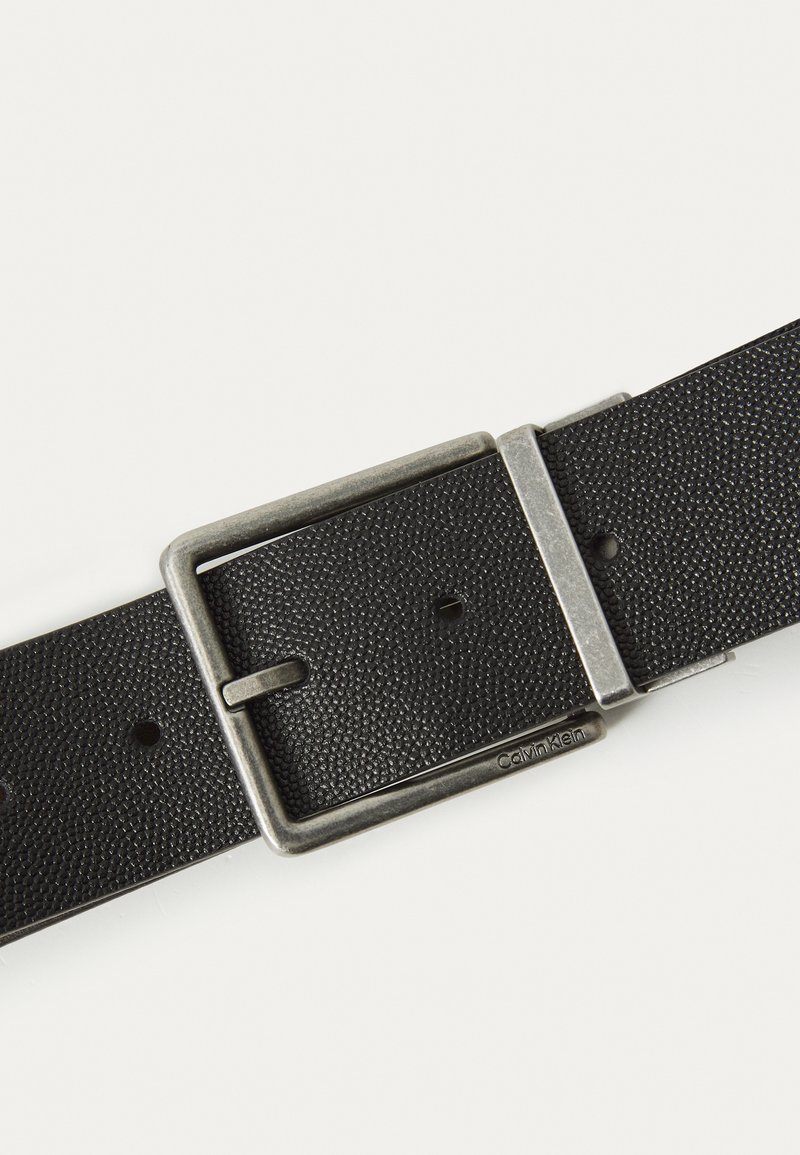 Schwarzer, strukturierten Leder Gürtel mit einer silbernen Metallschließe, die rechteckig geformt ist und mehrere Einstelllöcher aufweist. Mit "Calvin Klein" gekennzeichnet.