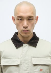 Νεαρός φαλακρός άντρας με σκουλαρίκια, φορώντας μπεζ σακάκι με σκούρο γιακά και μπροστινές τσέπες. Κοιτάει κατευθείαν στην κάμερα.