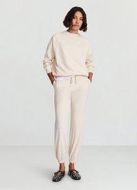 Sweat-shirt beige avec manches raglan et col rond, associé à un pantalon de jogging assorti avec taille et poignets élastiqués. Chaussures noires à enfiler avec un motif.