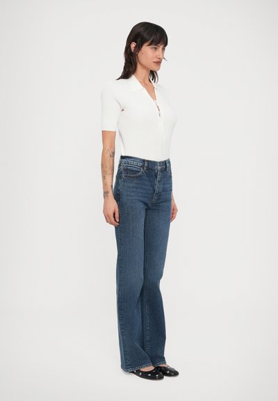 Vállig érő sötét hajú nő fehér rövid ujjú felsőben, magas derekú kék farmerral és fekete, ezüst szegecses lapos cipőben.