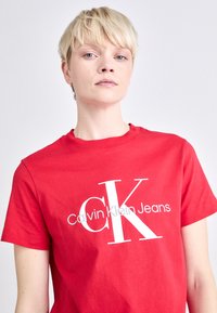 Rode katoenen T-shirt met een groot wit "CK" logo en daaronder de tekst "Calvin Klein Jeans" in een modern lettertype. Klassieke ronde halslijn en korte mouwen.