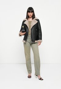Zwarte schapenvacht jas met een beige nepbont kraag, gecombineerd met lichtgroene jeans met versieringen en zwarte hakken. Houdt een zwarte handtas vast.