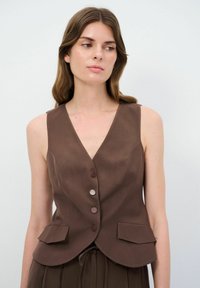 Chaleco marrón sin mangas con escote en V, que presenta una parte delantera con botones, dos bolsillos frontales y un diseño ajustado. Textura suave y forma entallada.