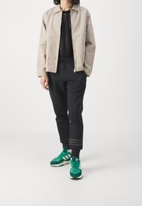 Giacca leggera beige, top nero testurizzato, pantaloni jogger neri con righe e sneakers verdi con accenti bianchi. Insieme casual.